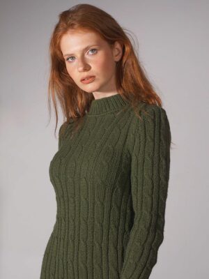 A model wearing a cable knit merino maxi dress in a soft green color, standing in front of a white background. The dress features a high neckline, long sleeves, and a fitted silhouette